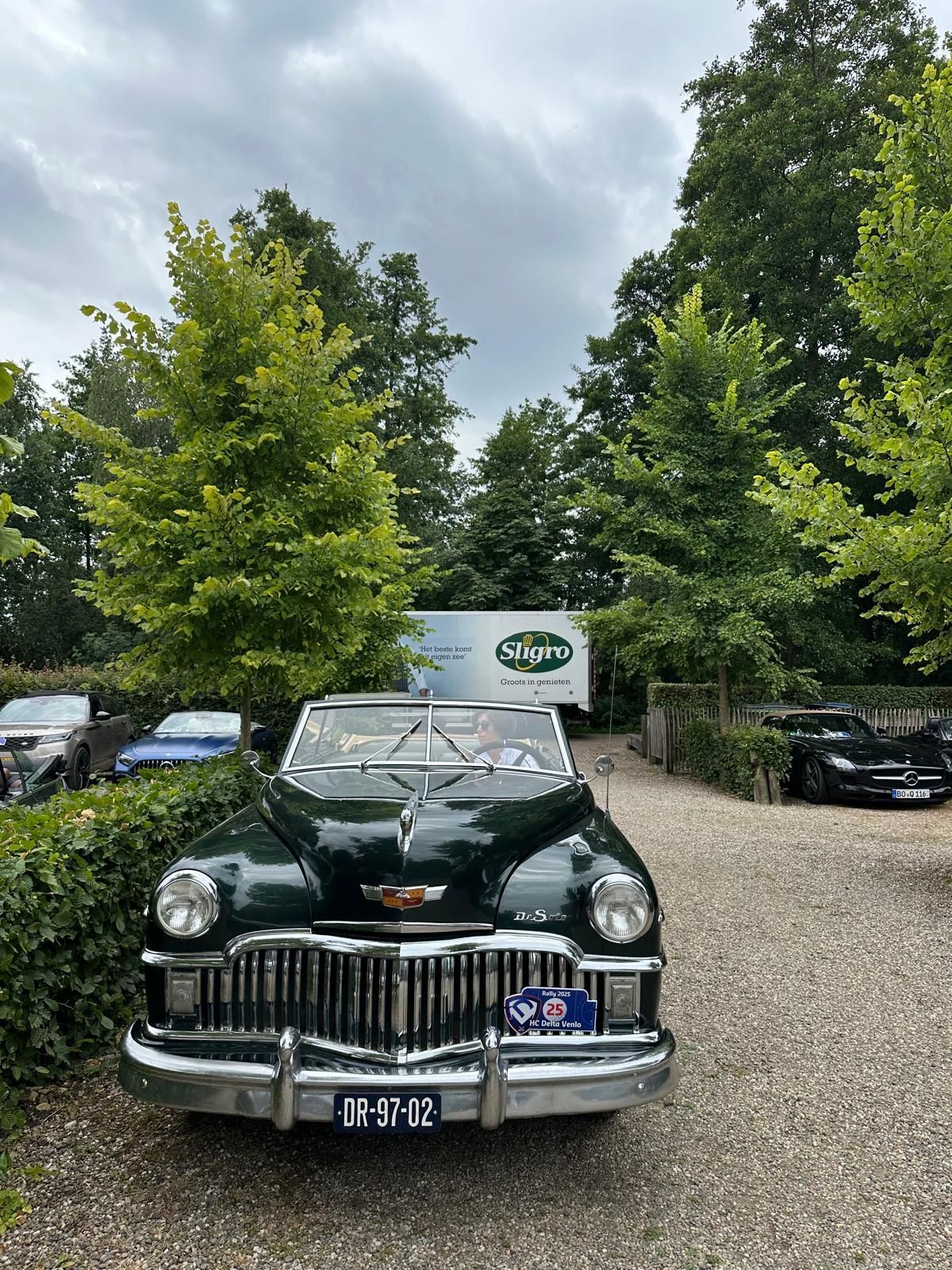 Donkergroene vintage cabriolet geparkeerd op een grindpad, bomen op de achtergrond, bewolkte lucht.