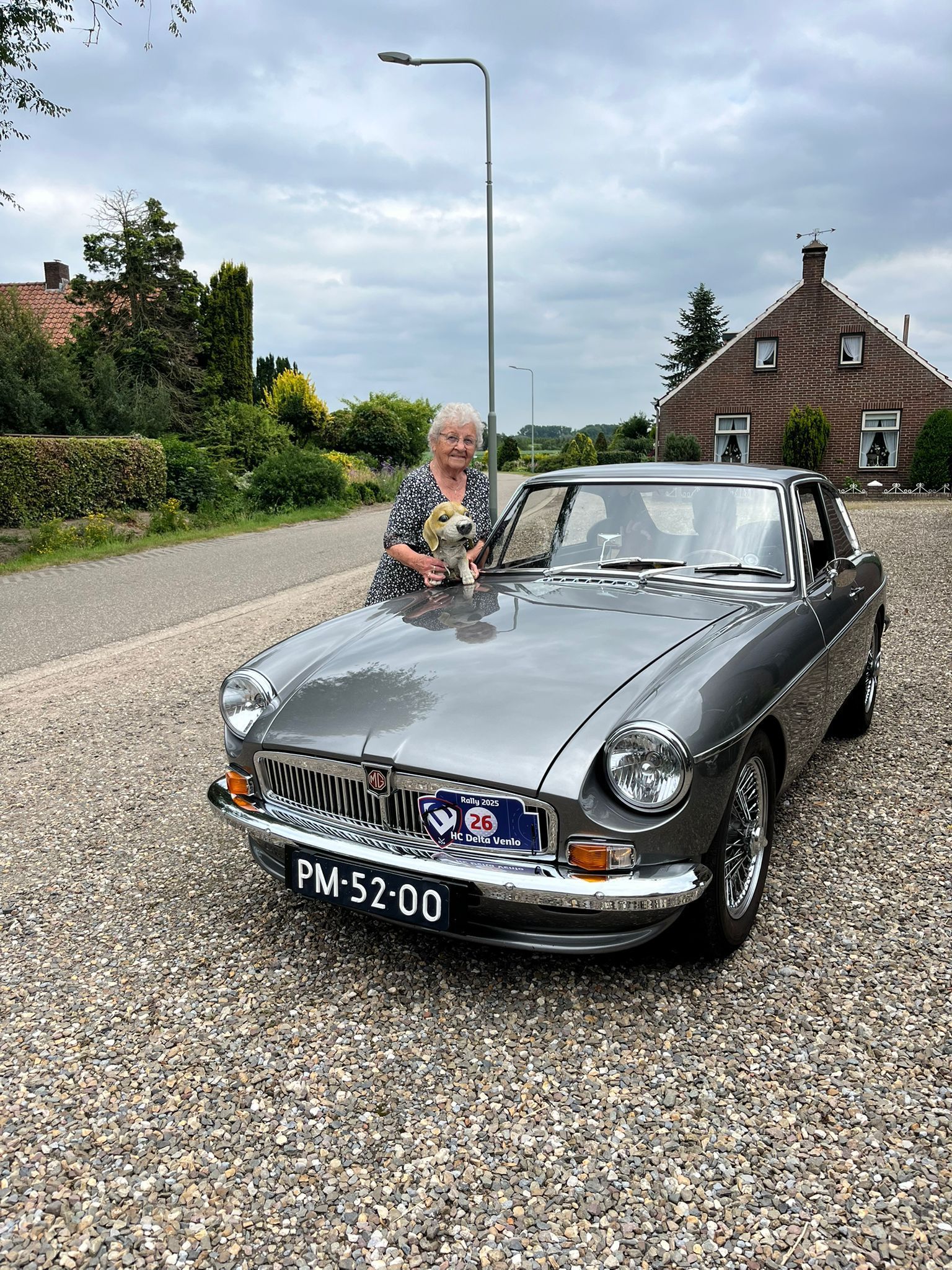 Een vrouw met een klein hondje poseert naast een grijze oldtimer op een grindpad. Op de achtergrond zijn een huis en bomen te zien.