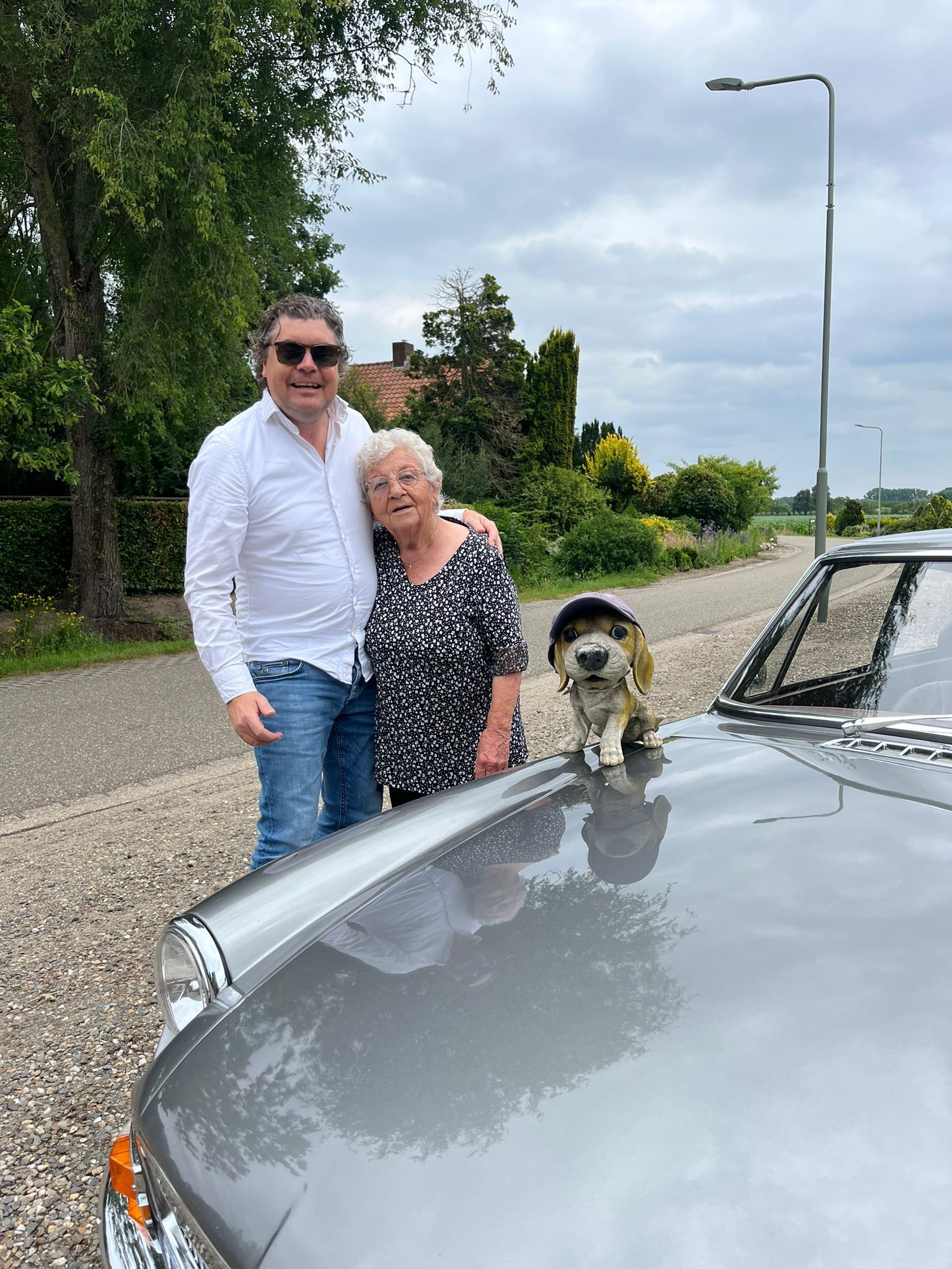 Een man en een vrouw staan naast een grijze auto, met een hond op de motorkap; de auto bevindt zich buiten en de lucht is bewolkt.