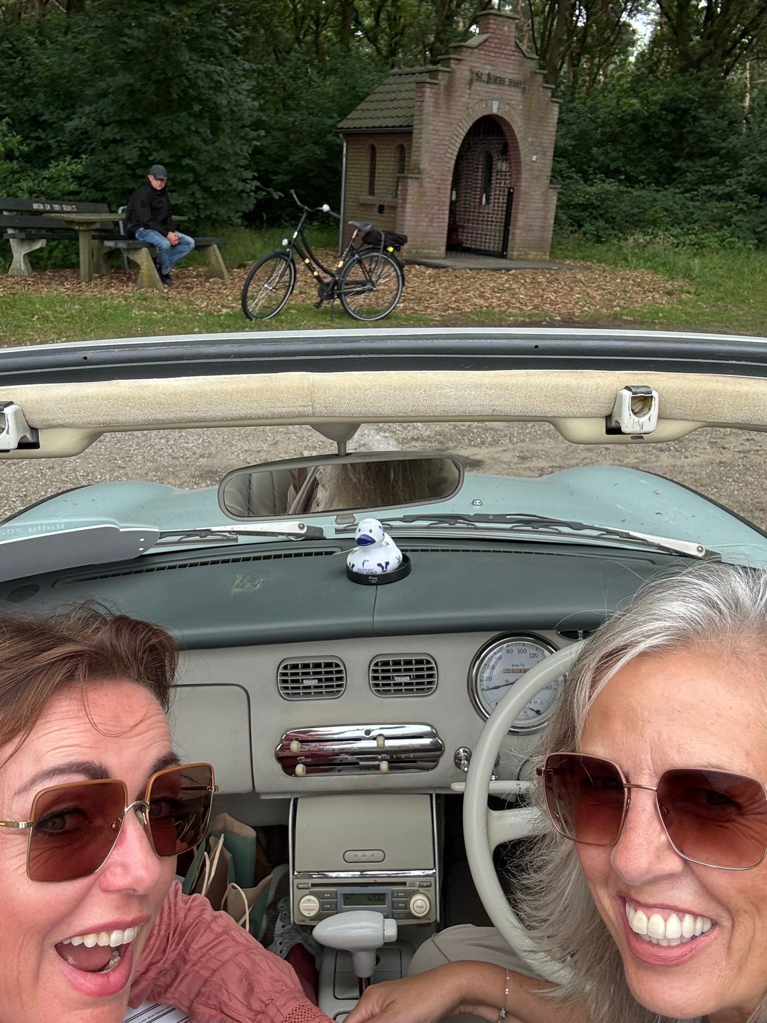 Twee vrouwen glimlachen in een oldtimer; op de achtergrond zijn een kapel, een fiets en een persoon op een bankje te zien.