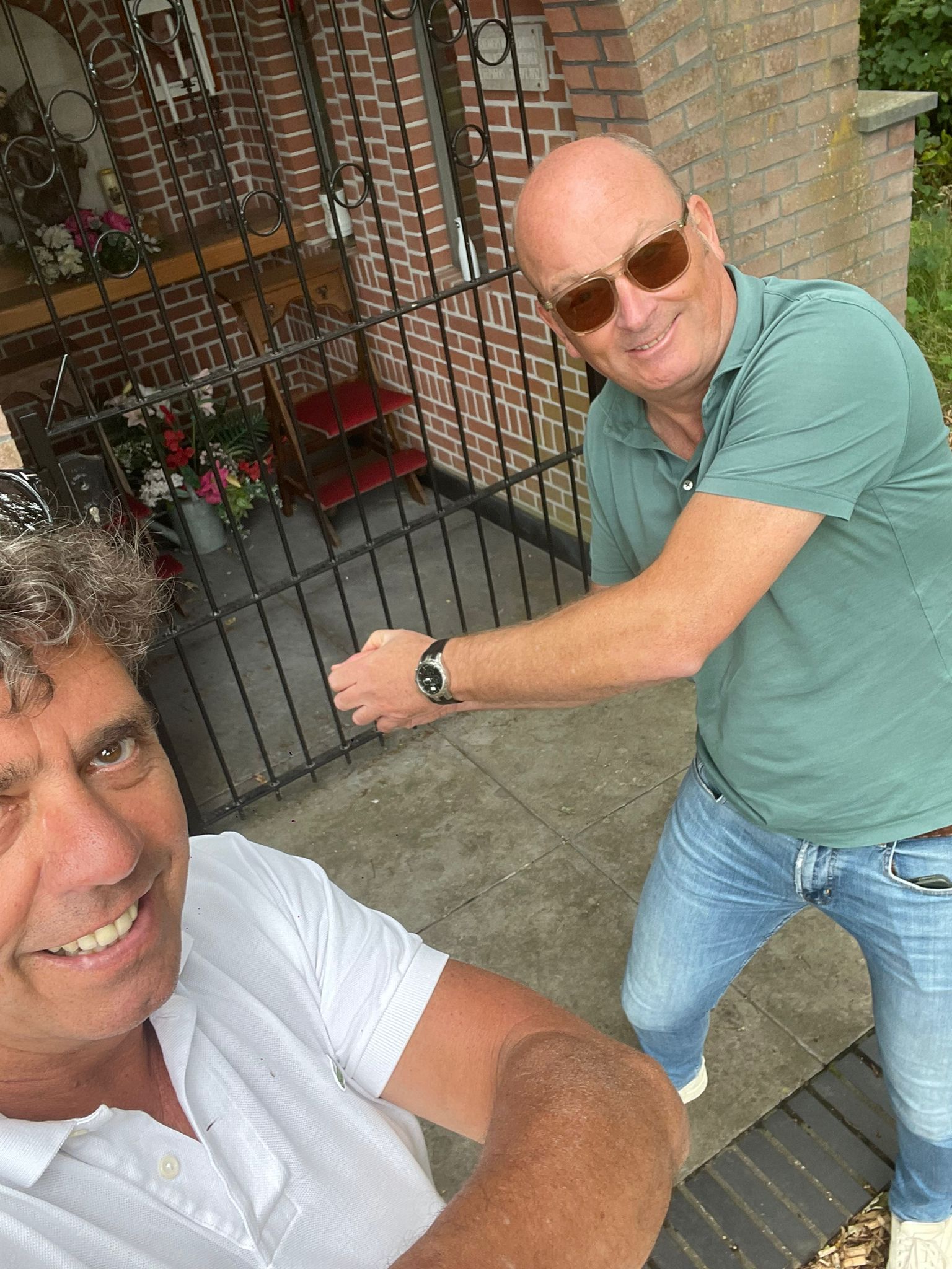 Twee mannen staan bij een bakstenen constructie met een poort. Een van hen wijst met uitgestrekte arm. Beiden glimlachen.