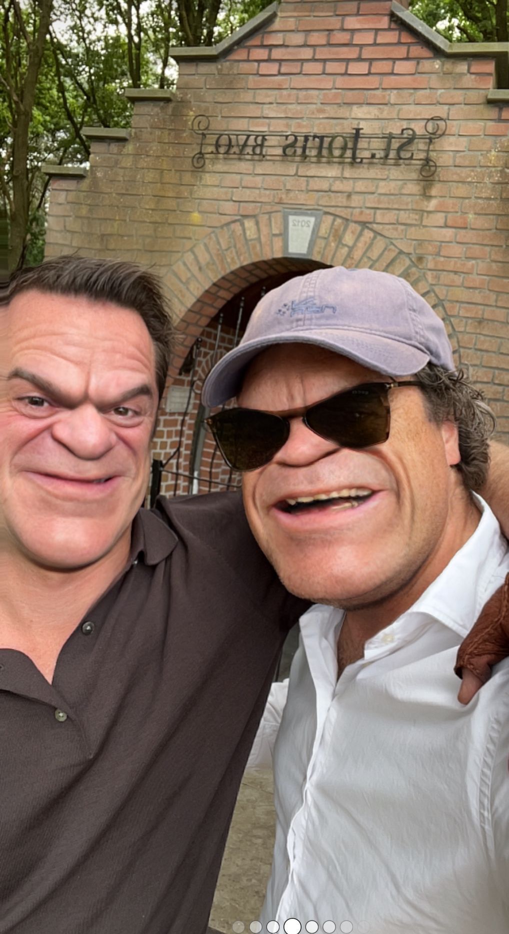 Twee mannen met overdreven gelaatstrekken poseren voor een selfie voor een bakstenen gebouw met een boogvormige ingang.