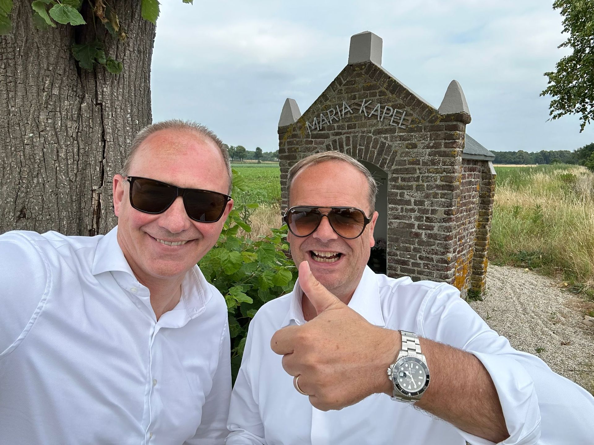 Twee mannen glimlachen en steken hun duim omhoog naast een kleine stenen kapel.
