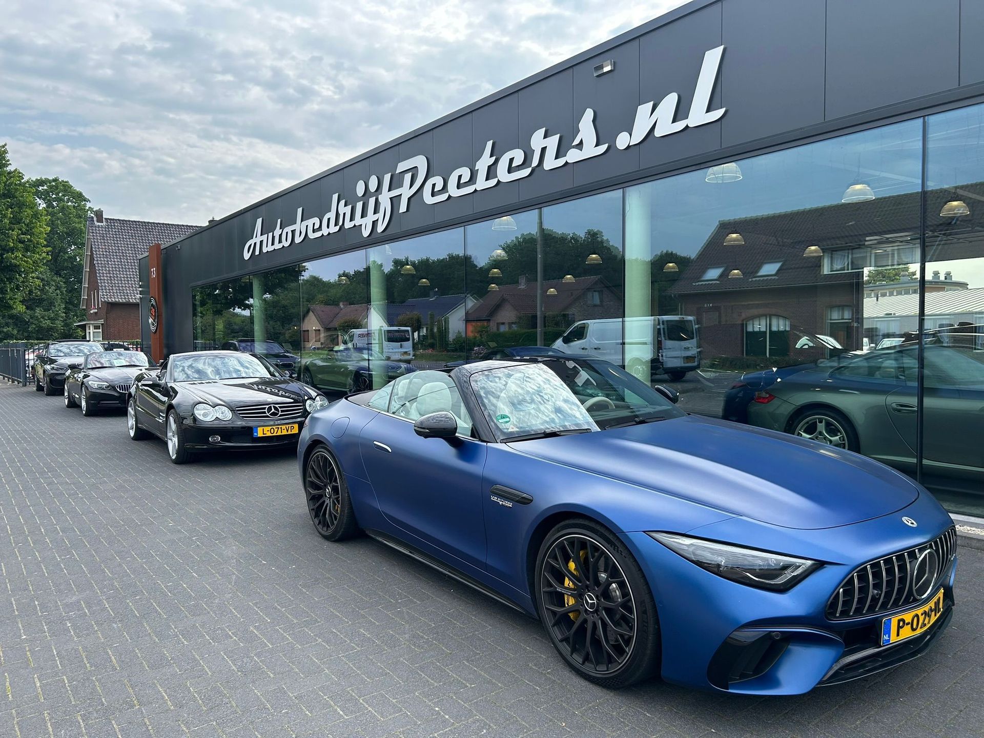 Een blauwe cabriolet Mercedes-Benz staat geparkeerd voor een autodealer, met andere voertuigen in de rij.