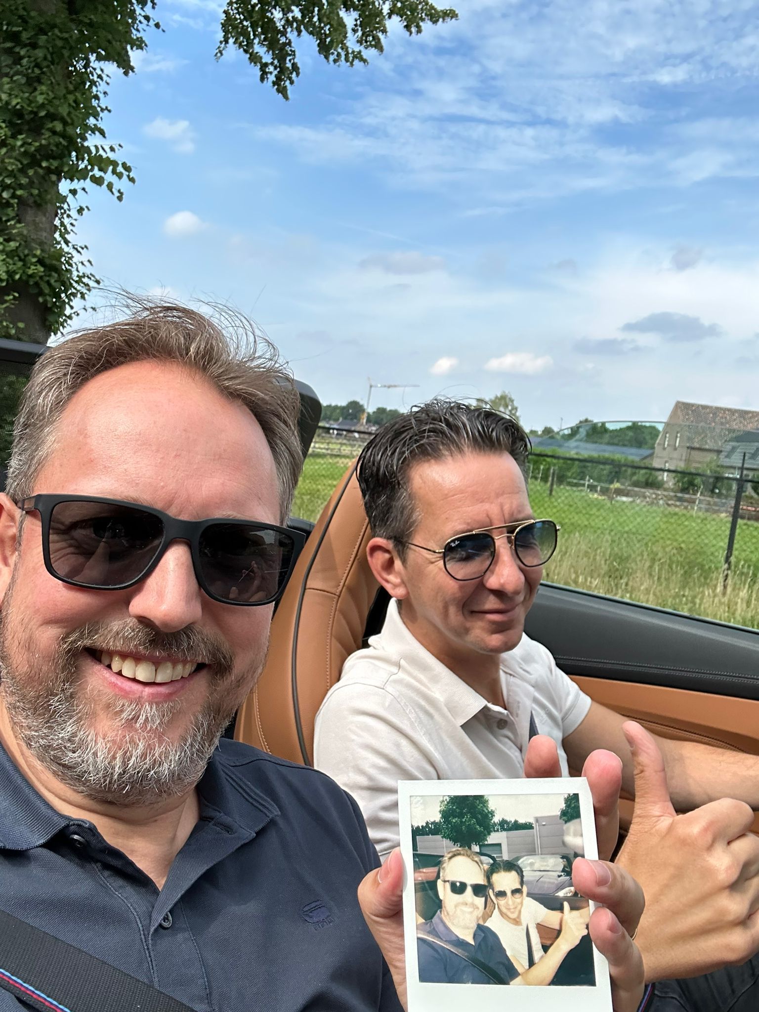 Twee mannen in een cabriolet, een van hen met een foto van zichzelf in de hand, lachend onder een blauwe hemel.