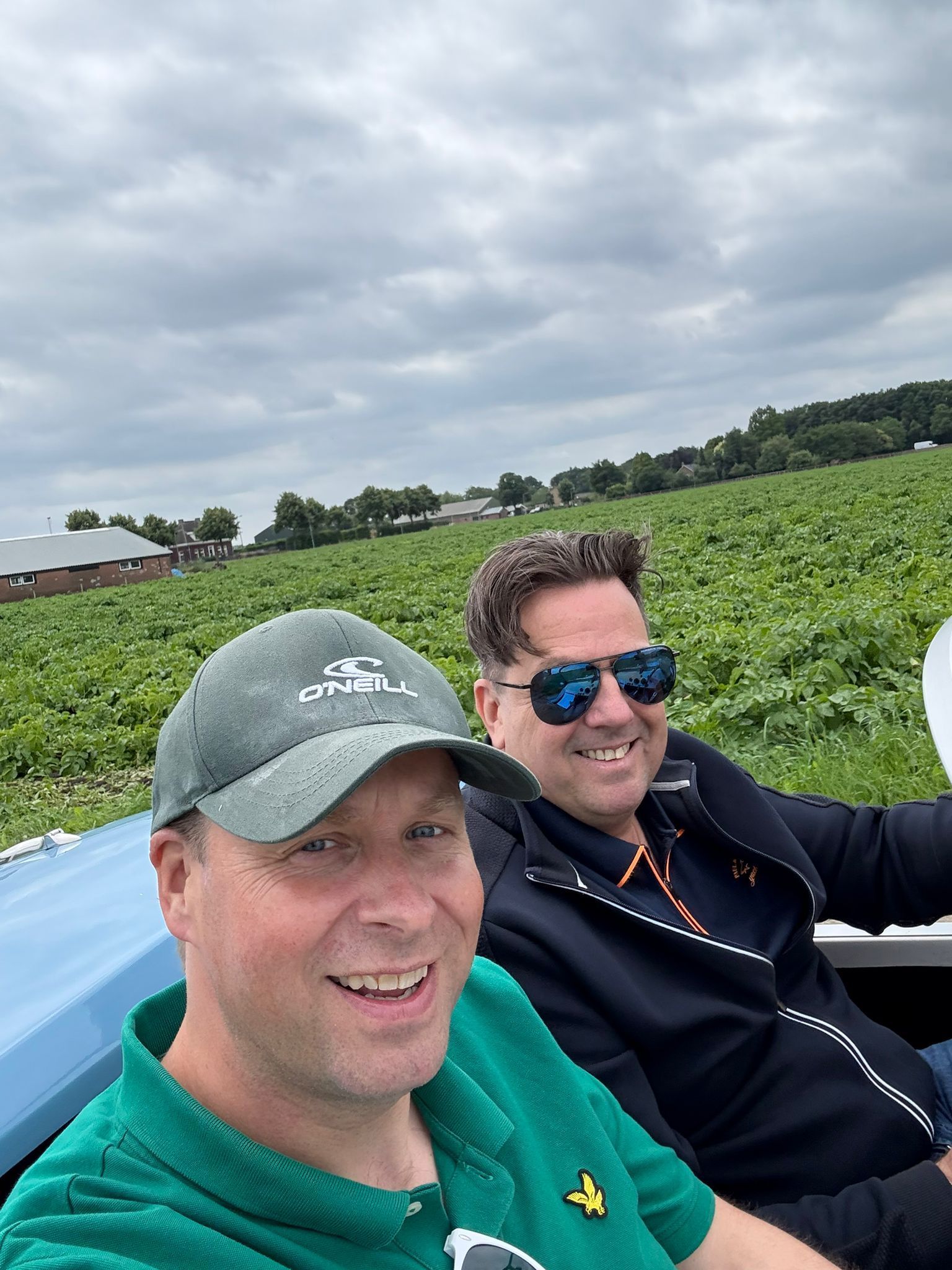 Twee mannen staan lachend in een veld. De ene draagt een groene pet, de andere een zonnebril. Boven hen is een bewolkte hemel.