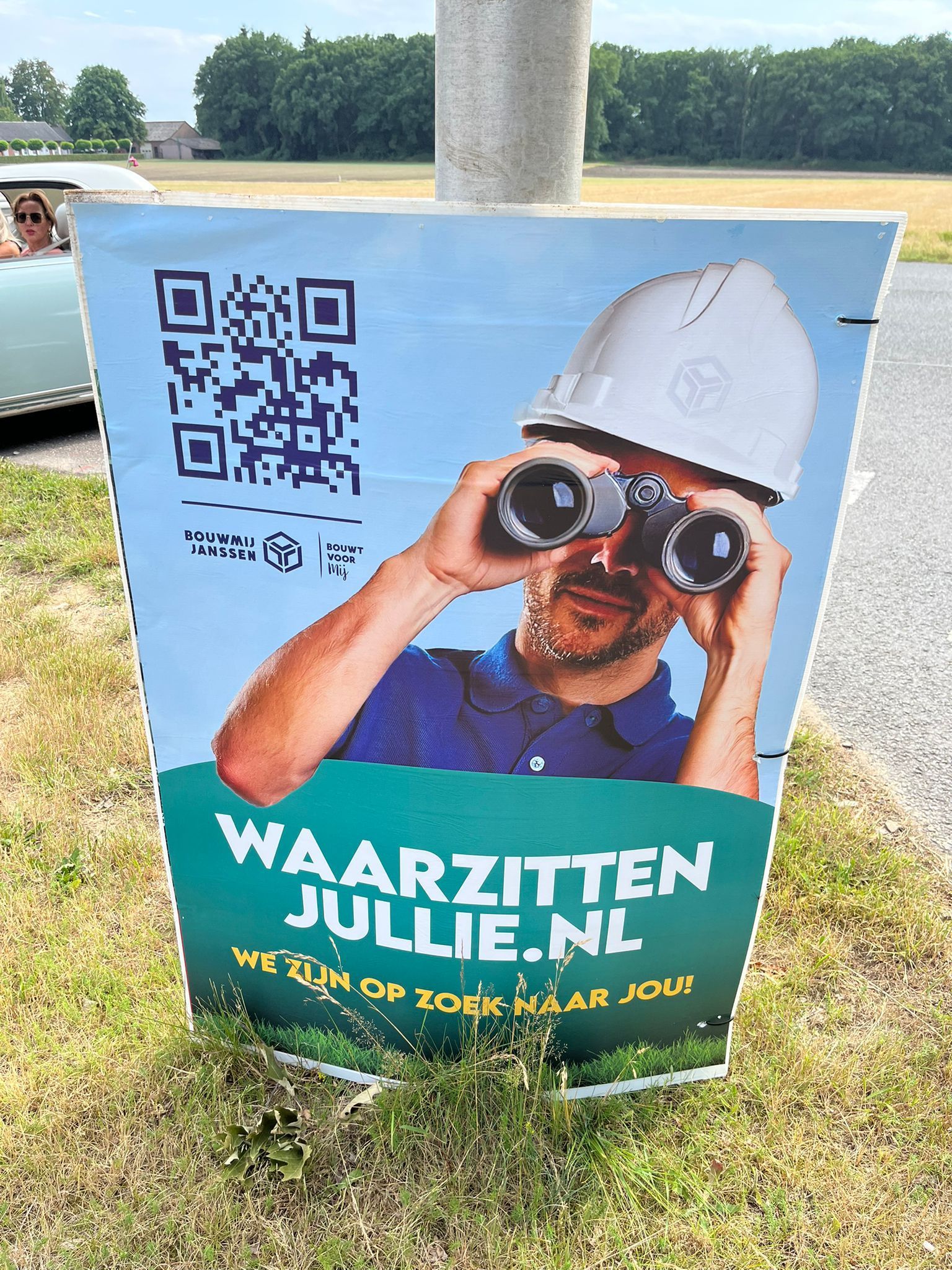 Bord met een man met een helm die door een verrekijker kijkt, websiteadres en QR-code.