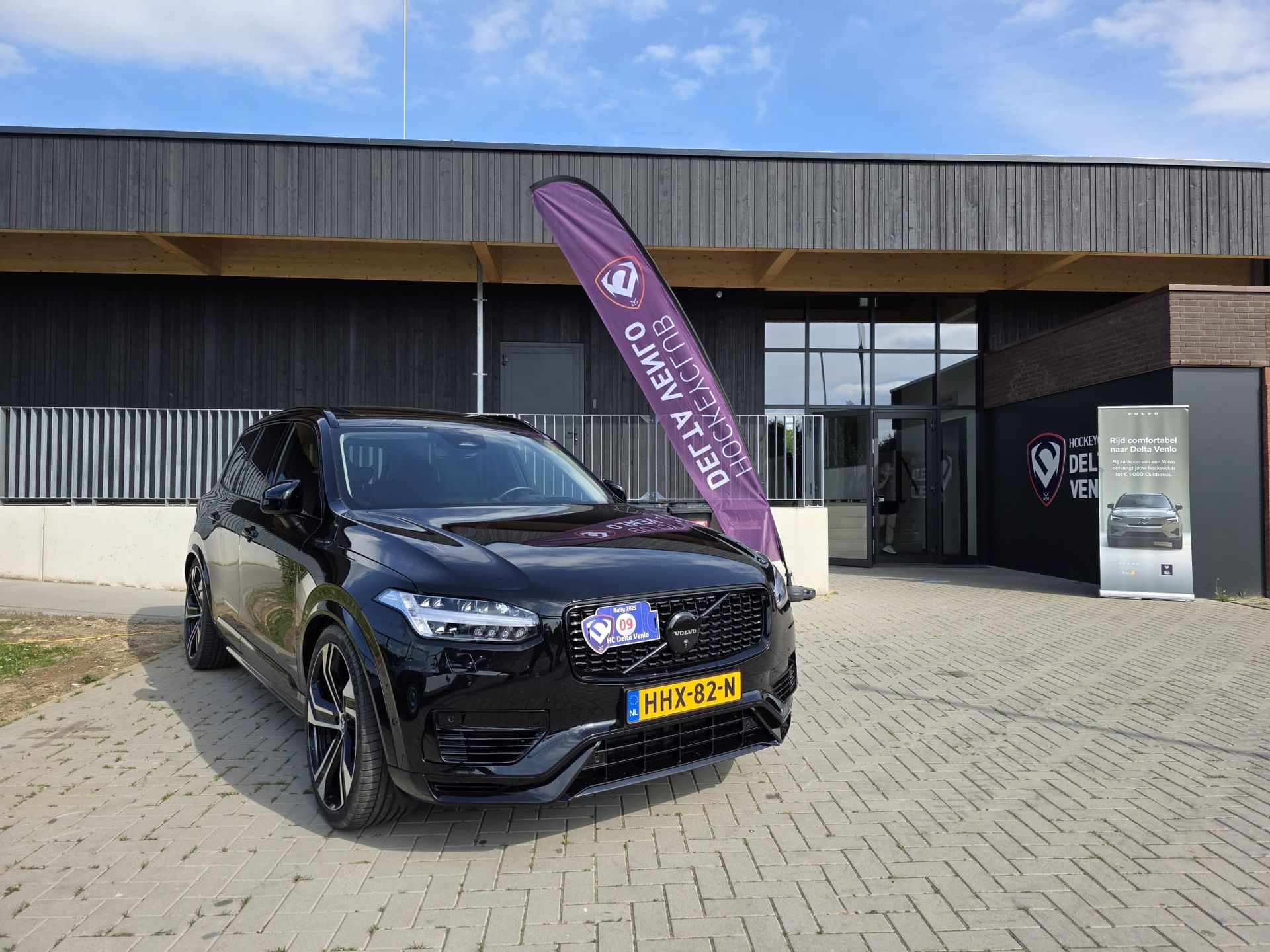 Een zwarte SUV staat geparkeerd voor een donker gebouw met een bord en een paarse vlag op een zonnige dag.