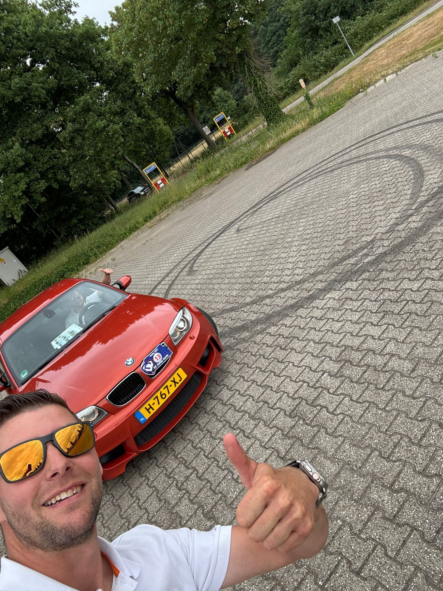 Een man steekt zijn duim omhoog naast een rode BMW, met bandensporen op het wegdek.