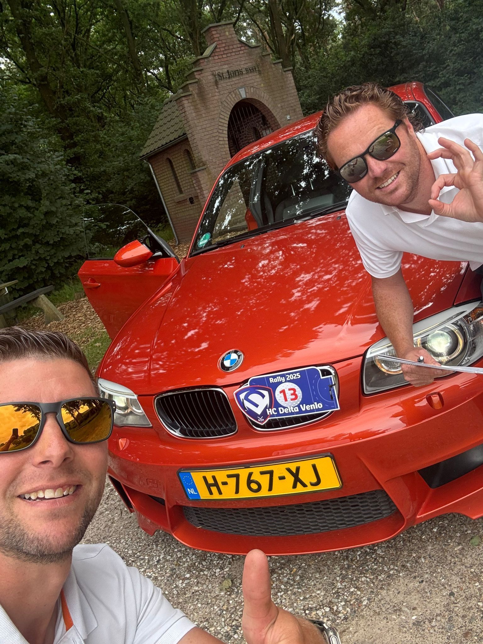 Twee mannen poseren bij een feloranje BMW. De ene steekt zijn duim omhoog, de andere maakt een
