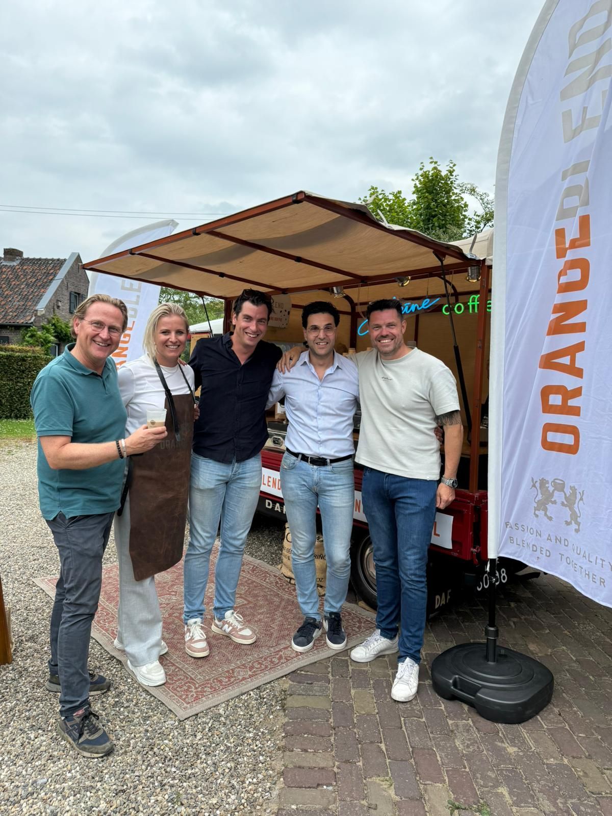 Vijf mensen poseren bij een voedselkar met oranje logo; twee mensen hebben hun armen om elkaar heen geslagen, buiten.