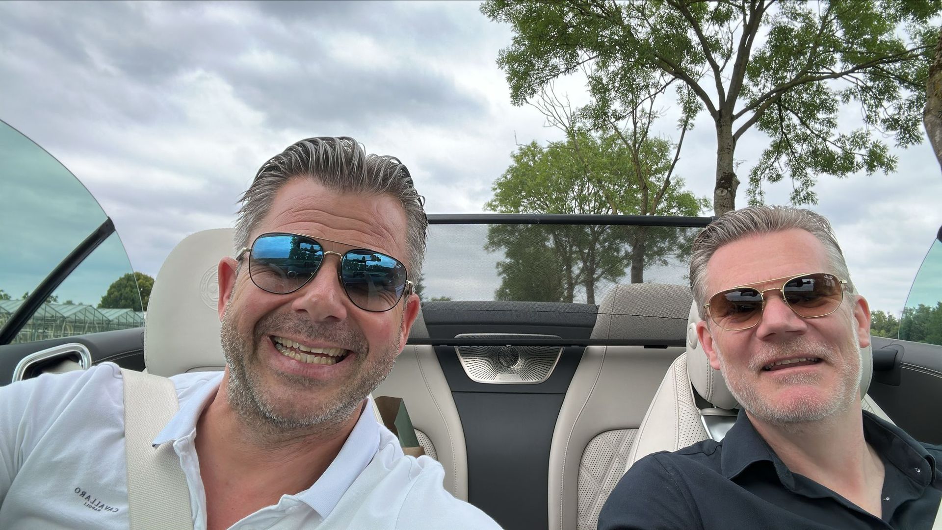 Twee mannen in een cabriolet, met zonnebrillen op, lachend, rijdend op een bewolkte dag.