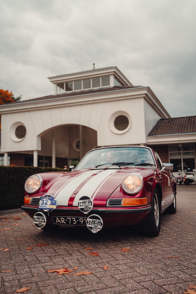Een klassieke Porsche met rode en witte strepen staat geparkeerd voor een wit gebouw met ovale ramen.
