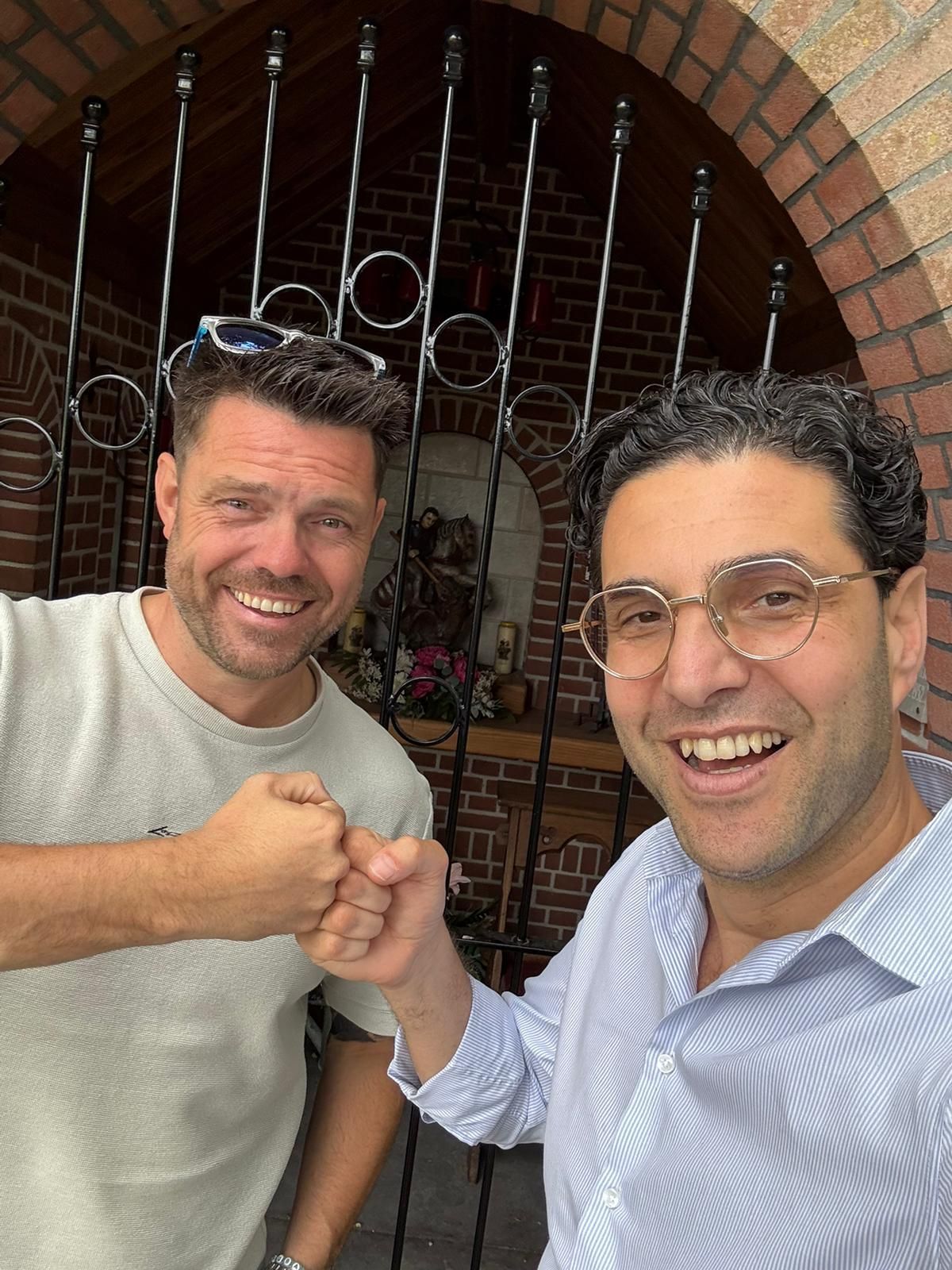 Twee mannen glimlachen en geven elkaar een vuistje voor een bakstenen muur met een sierlijke poort.