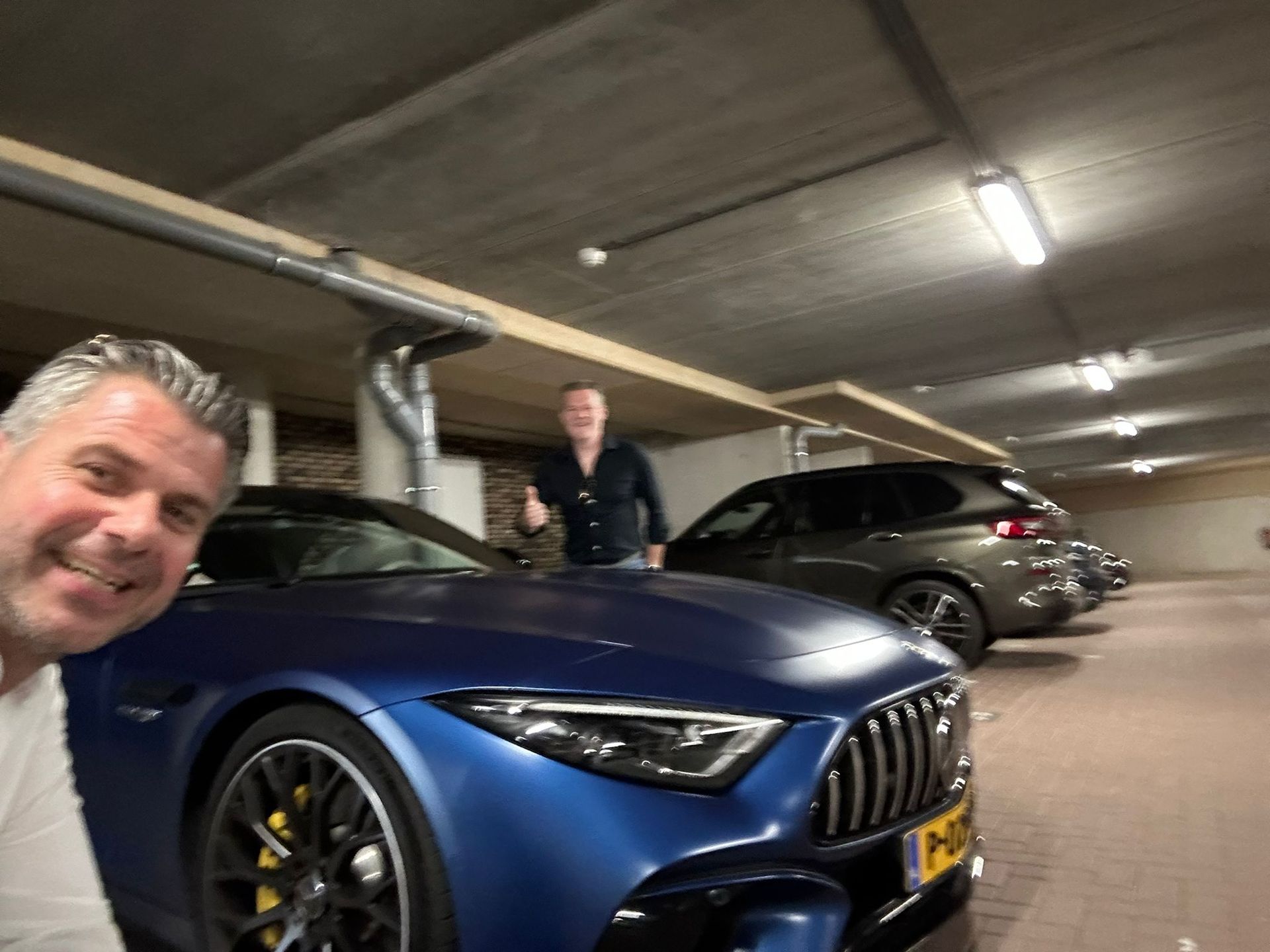 Twee mannen poseren met een blauwe sportwagen in een overdekte parkeergarage; de ene man lacht terwijl de andere zijn duim omhoog steekt.