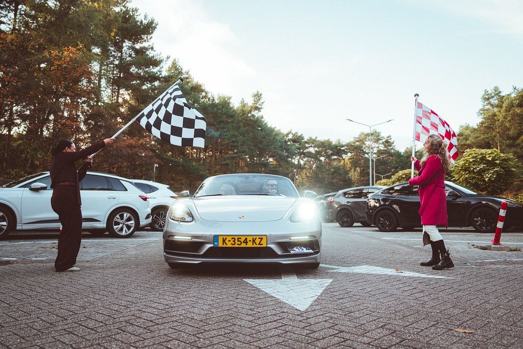 Een zilverkleurige sportwagen passeert de finishlijn, met vlaggen van twee mensen. Op de achtergrond zijn bomen en auto's te zien.