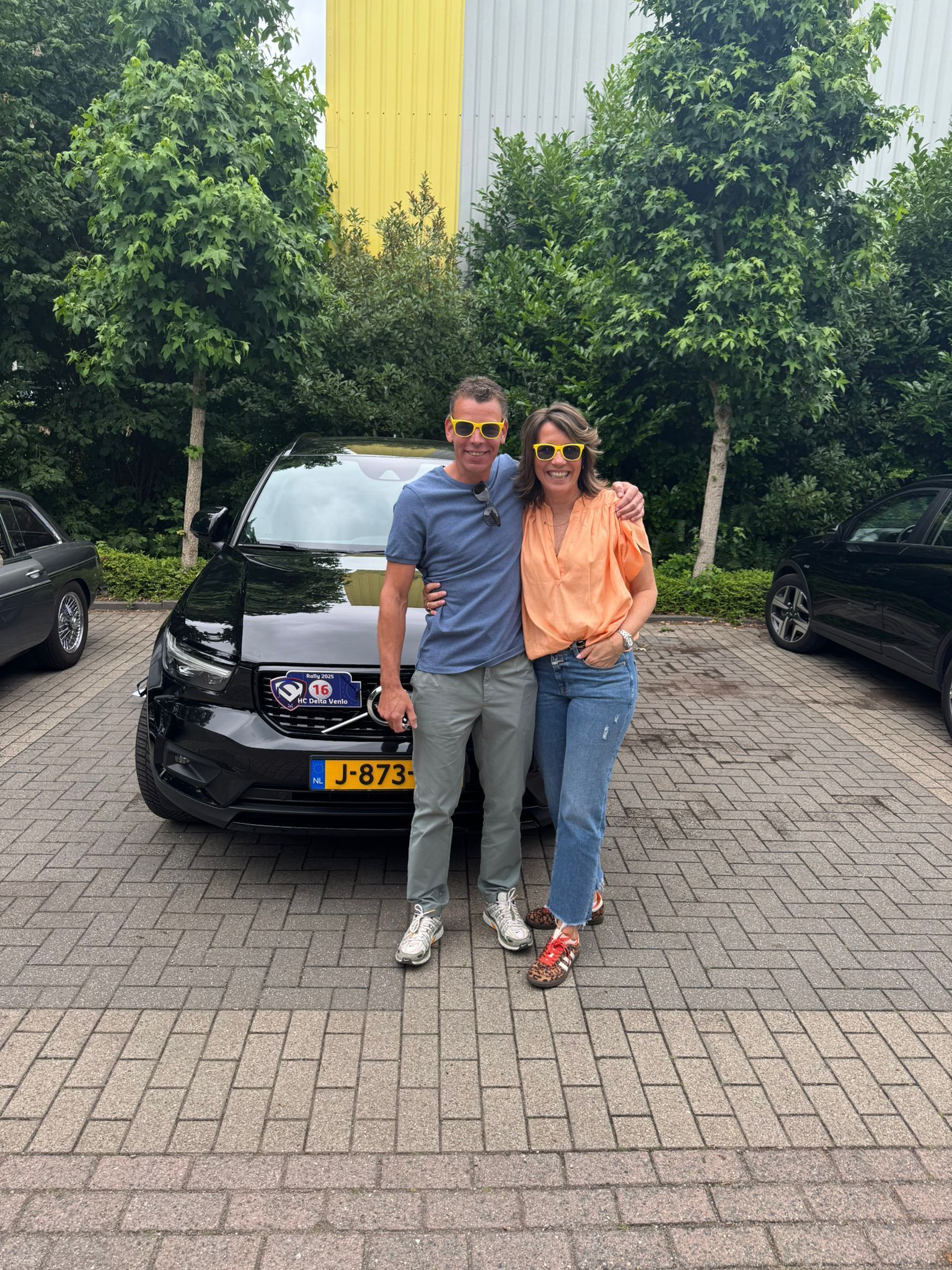 Twee mensen met zonnebrillen poseren voor een zwarte auto, met bomen op de achtergrond en een gebouw in de verte.
