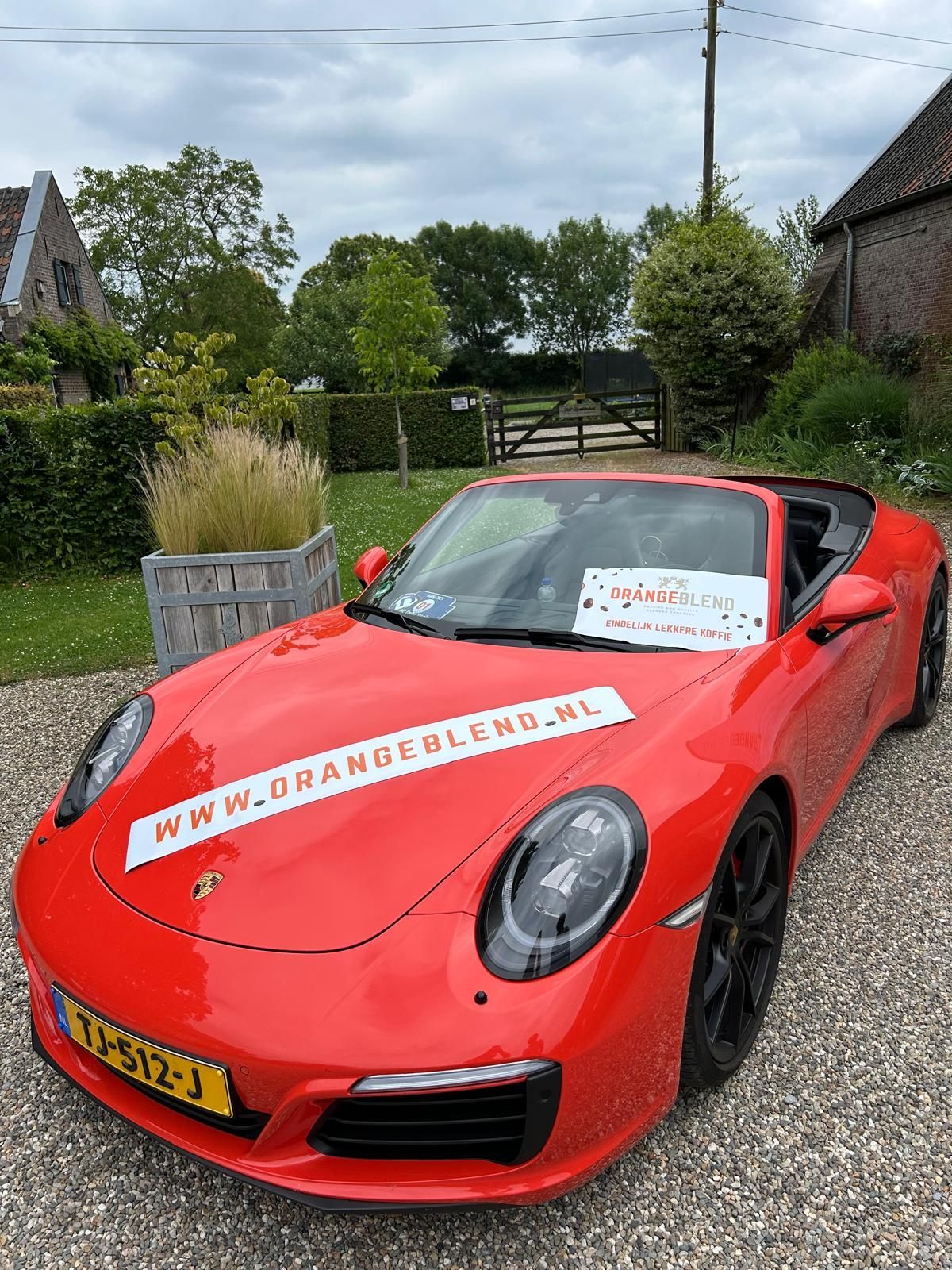 Een rode Porsche cabriolet staat geparkeerd op een grindpad, met een websitebanner op de motorkap.