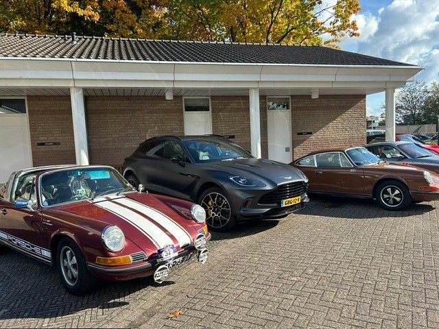 Er stonden meerdere auto's geparkeerd voor een gebouw: een klassieke bordeauxrode Porsche, een grijze SUV en een bruine Porsche.