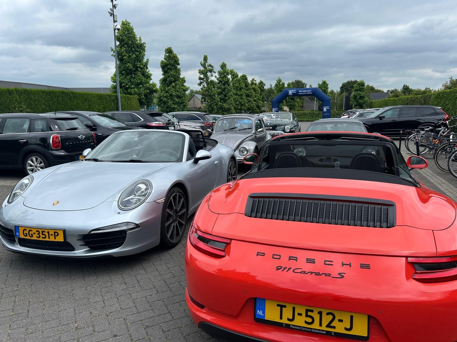 Twee Porsche cabriolets, een zilveren en een rode, staan geparkeerd op een parkeerplaats tussen andere auto's onder een bewolkte hemel.