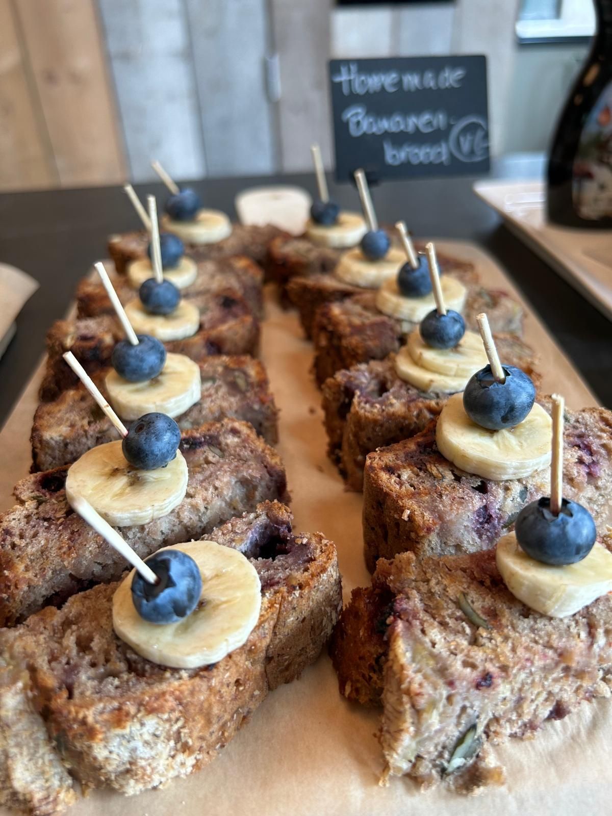 Plakjes zelfgebakken bananenbrood, belegd met plakjes banaan en bosbessen, op een houten dienblad.