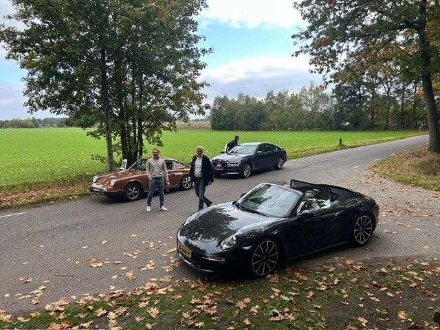 Drie mannen staan ​​bij klassieke auto's op een weg, met een veld op de achtergrond.