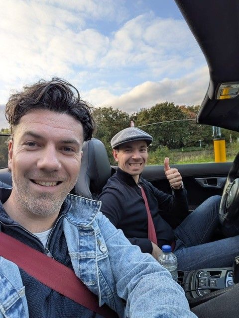 Twee mannen in een cabriolet, lachend. Een van hen steekt zijn duim omhoog. Bewolkte lucht.