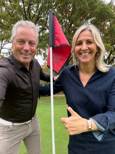 Man en vrouw bij een golfvlag; de vrouw steekt haar duim omhoog.