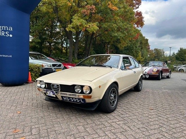 Een beige vintage Alfa Romeo coupé staat geparkeerd op een bakstenen weg tussen andere klassieke auto's in een herfstlandschap.