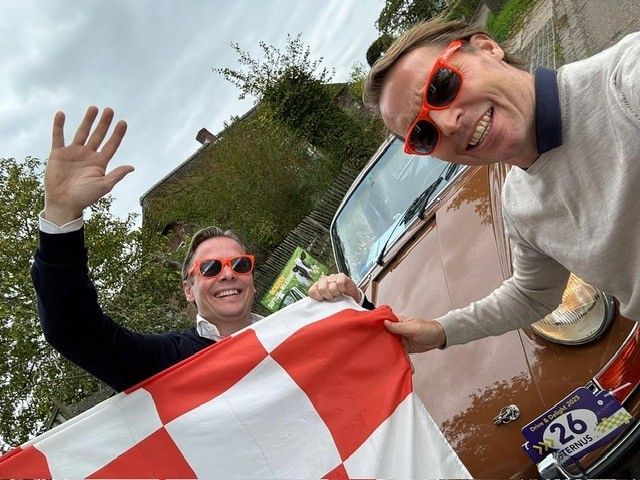 Twee mannen zwaaien met een geblokte vlag naast een oude bruine auto; ze dragen rode zonnebrillen.