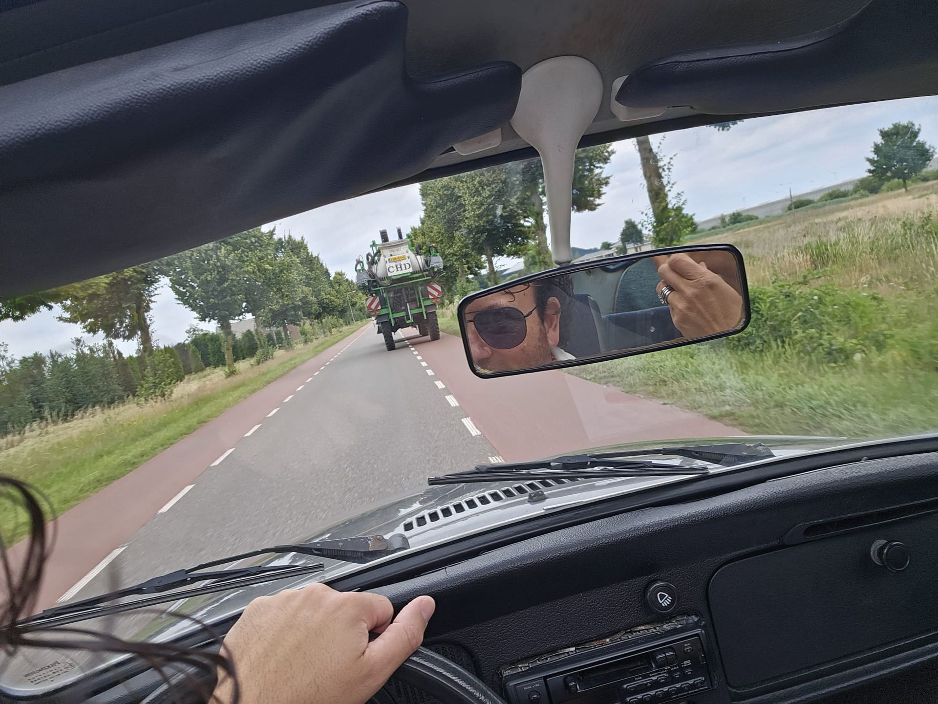 Een persoon die in een oldtimer rijdt, ziet een tractor in de achteruitkijkspiegel op een landweg.
