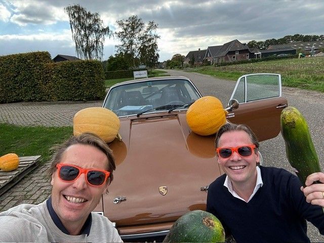 Twee mannen met oranje zonnebrillen poseren met pompoenen en een courgette op een vintage bruine auto. Landelijke buitenomgeving.