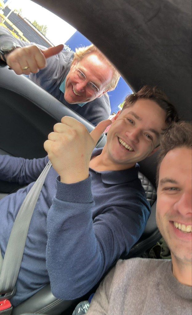 Drie mannen in een auto, lachend en met hun duimen omhoog.