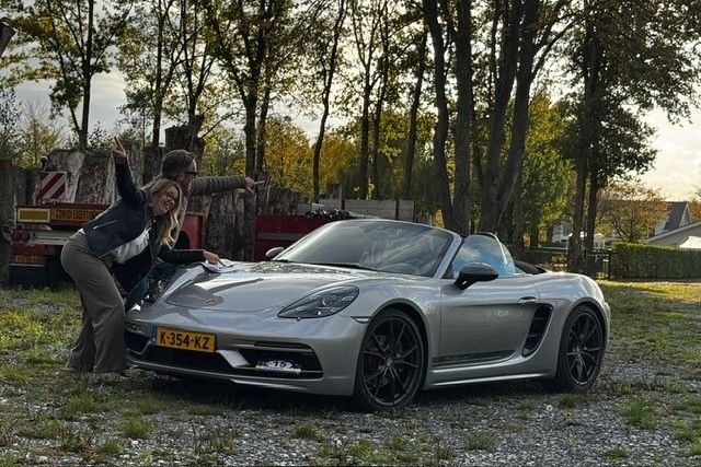 Twee mensen bewonderen een zilveren Porsche Boxster cabriolet in de buitenlucht, met bomen op de achtergrond.