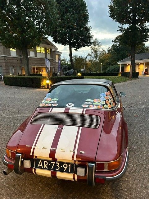 Een vintage Porsche 911 Targa in bordeauxrood met witte strepen, Nederlands kenteken, geparkeerd bij een gebouw en bomen.