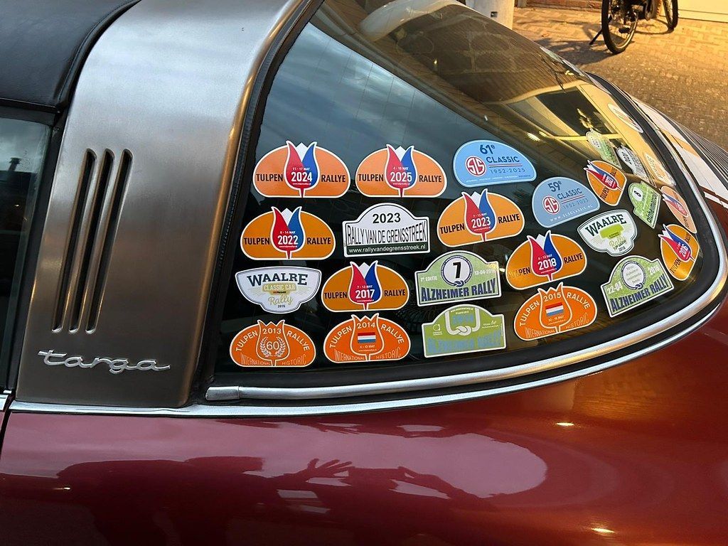 Close-up van de achterruit van een bordeauxrode Porsche Targa, beplakt met evenementstickers; op de achtergrond is een zwarte motorfiets te zien.
