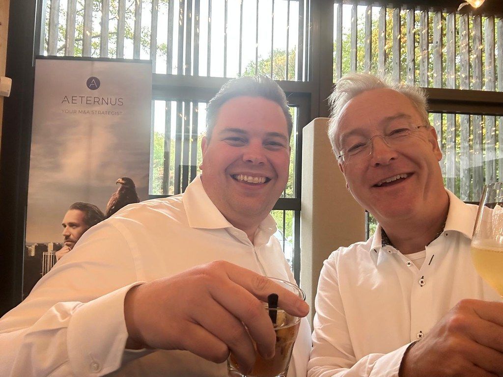 Twee mannen glimlachen en heffen hun glas. De ene houdt een donkere drank vast, de andere een heldere. Op de achtergrond is een bord te zien.