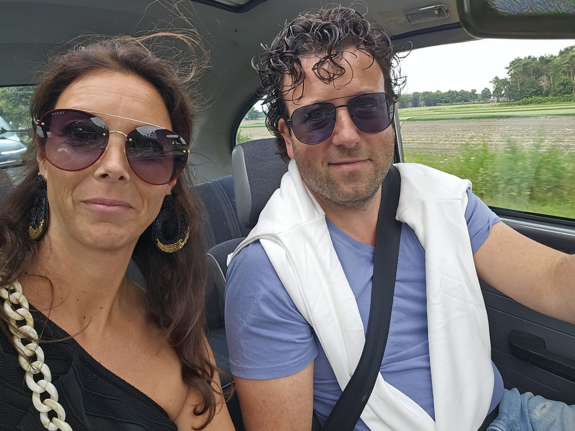 Twee mensen in een auto, beiden met een zonnebril op. De bestuurder heeft een handdoek over zijn schouders, de passagier draagt een grote bril.