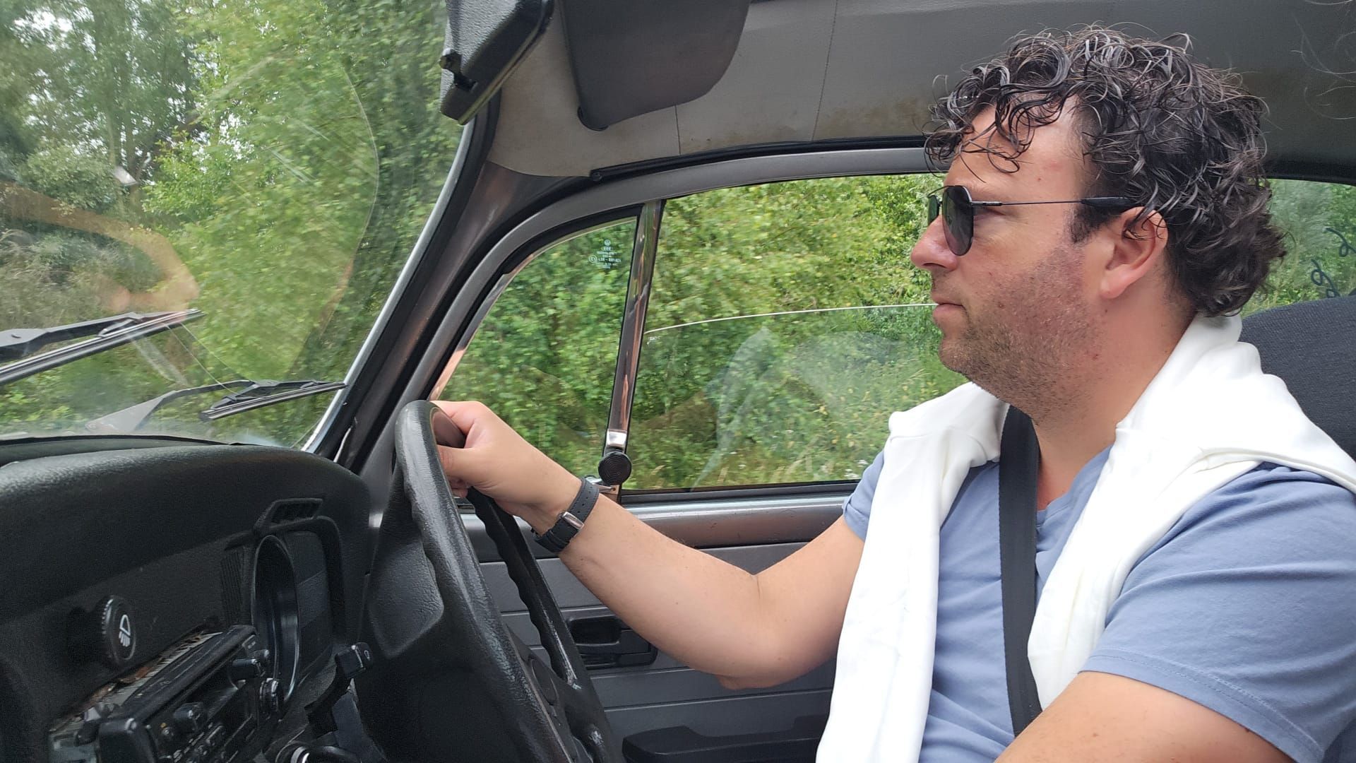 Een man rijdt in een oldtimer over een met bomen omzoomde weg, met een zonnebril en een witte trui aan.