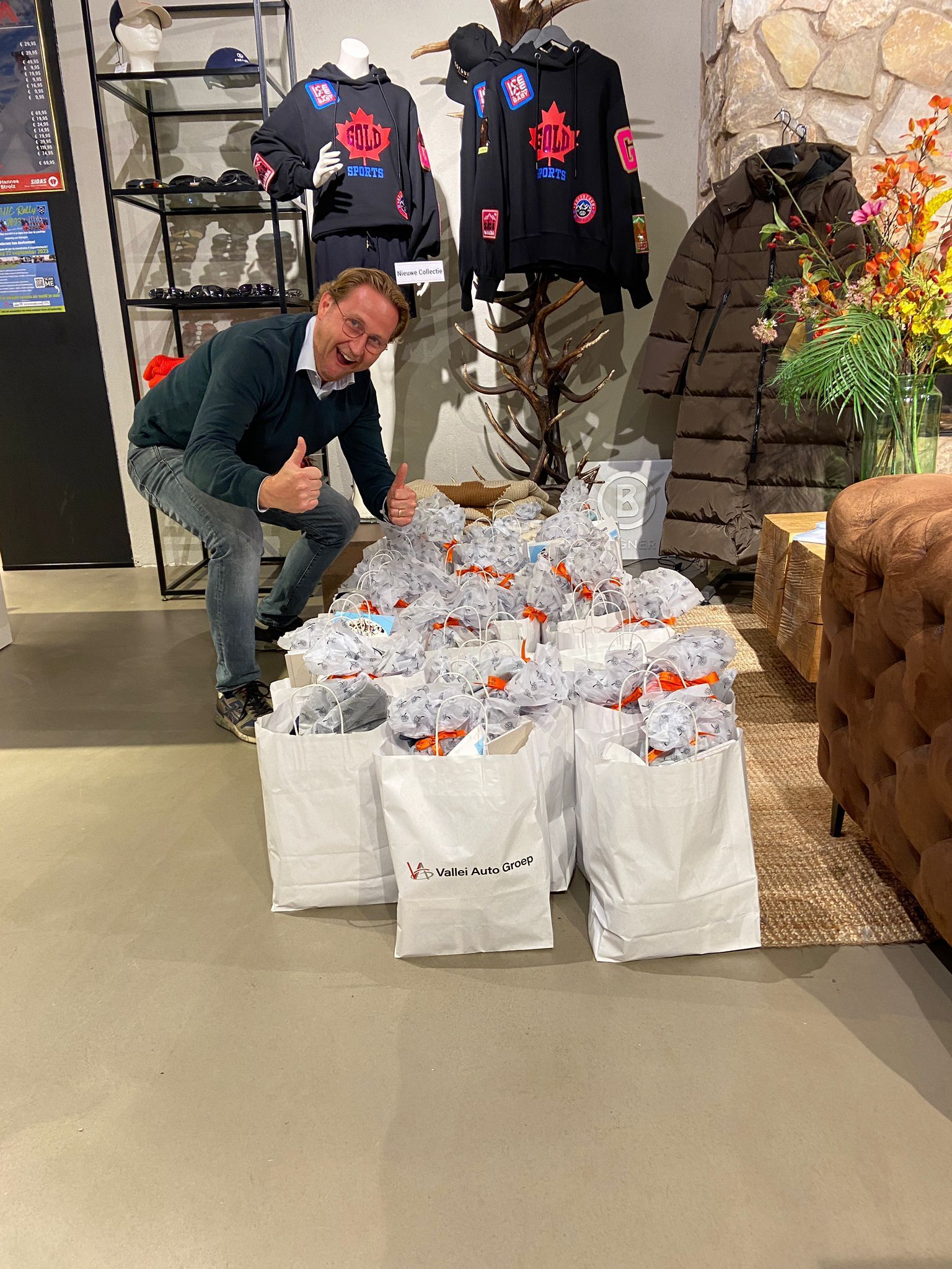 Een lachende man met een opgestoken duim staat naast een stapel cadeautassen in een winkel; op de achtergrond zijn kledingrekken te zien.