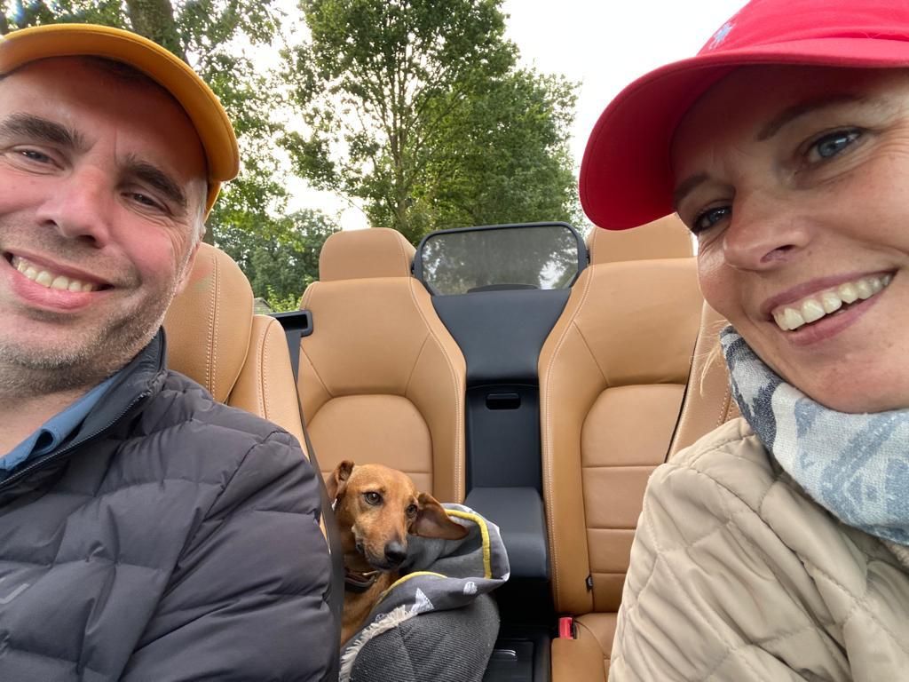 Twee mensen en een hond in een cabriolet. De man met de gele hoed lacht. De vrouw met de rode hoed lacht. De hond draagt ​​een jas.