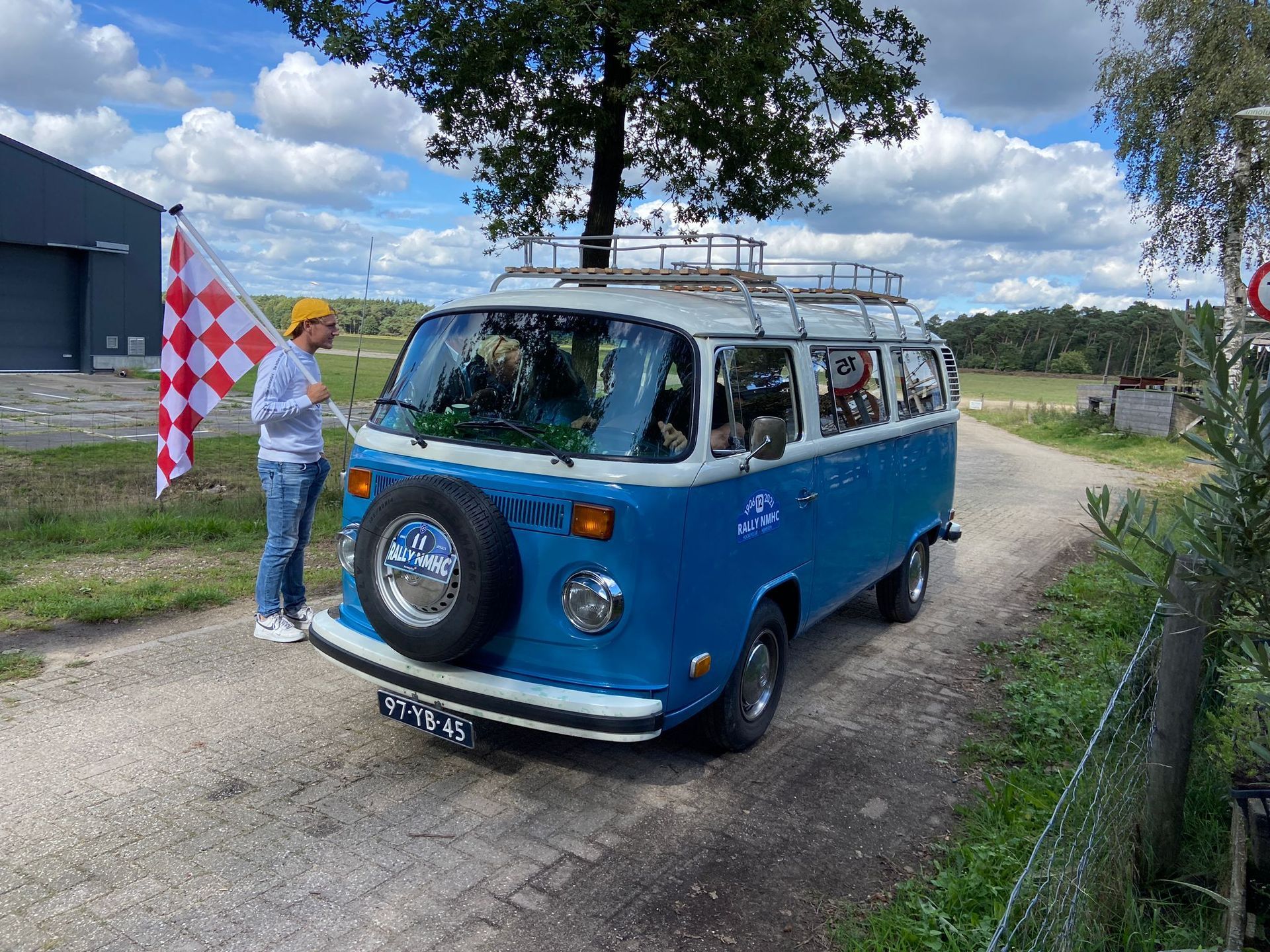 Een blauwe VW-bus met een reserveband, een persoon met een geblokte vlag, staand op een grindweg.