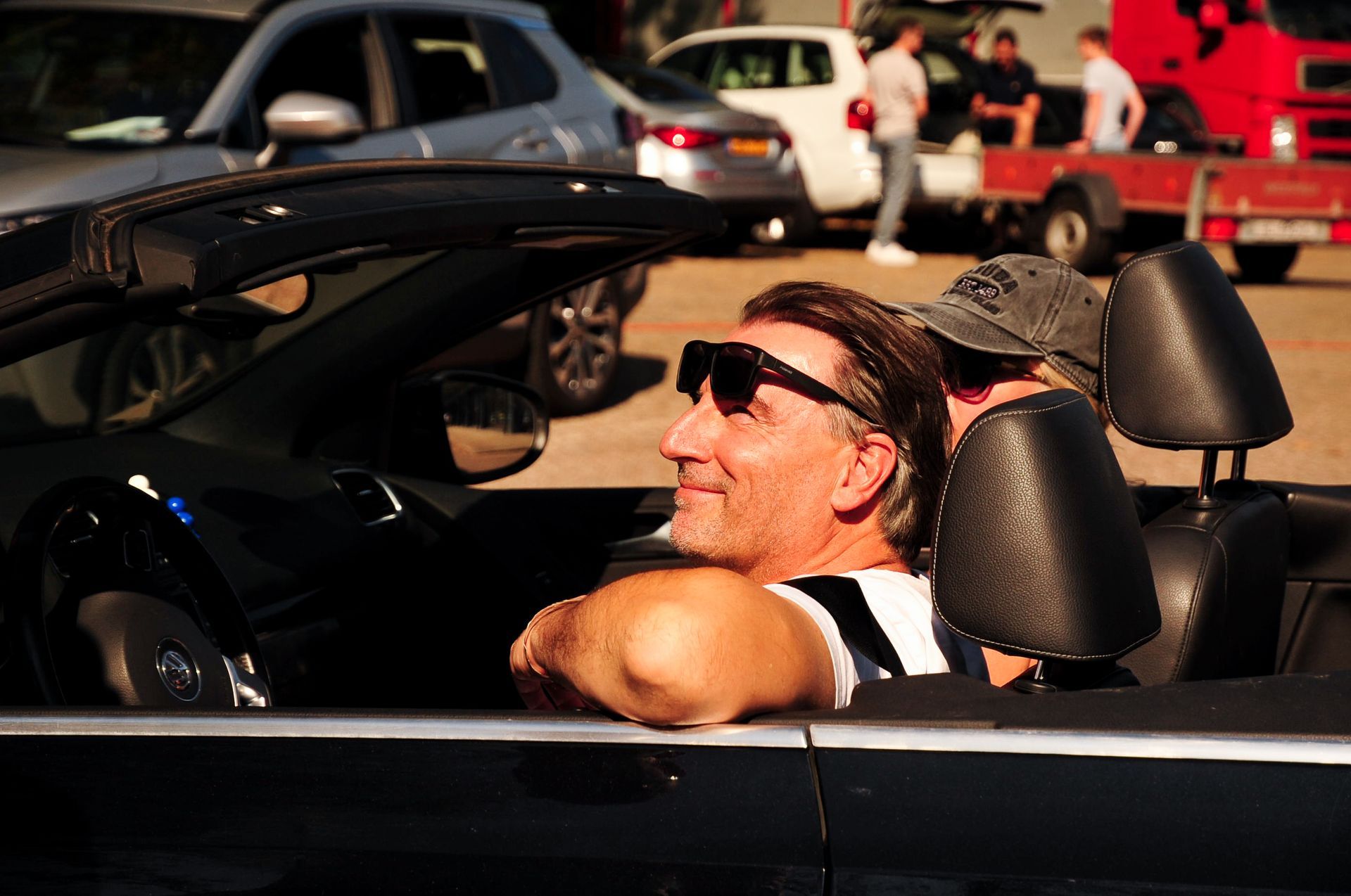 Een man met een zonnebril lacht in een zwarte cabriolet; op de achtergrond zijn andere voertuigen en mensen te zien.