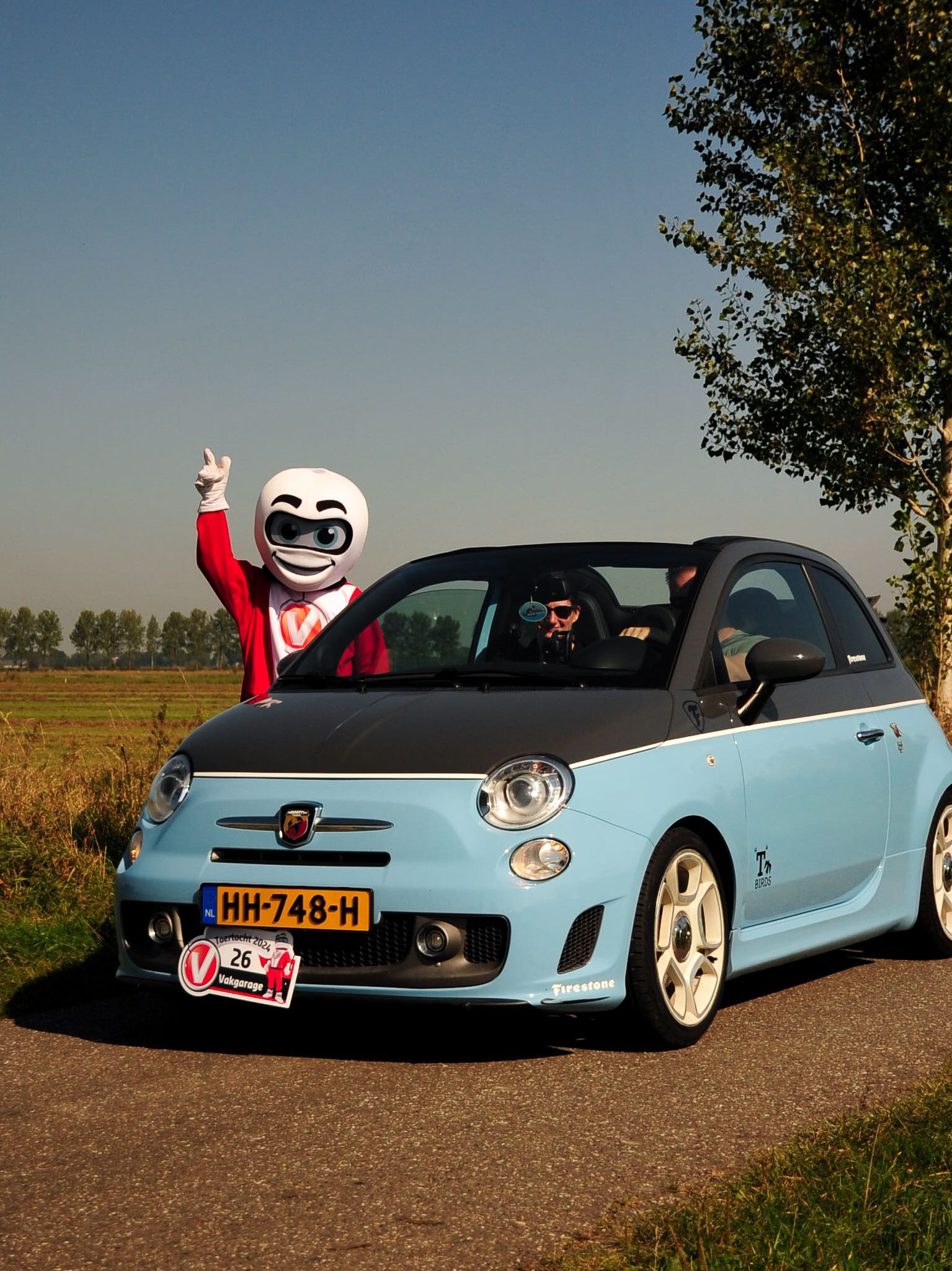 Een lichtblauwe Fiat 500 cabriolet met een verklede persoon met zonnebril die zwaait in de buurt van een veld.