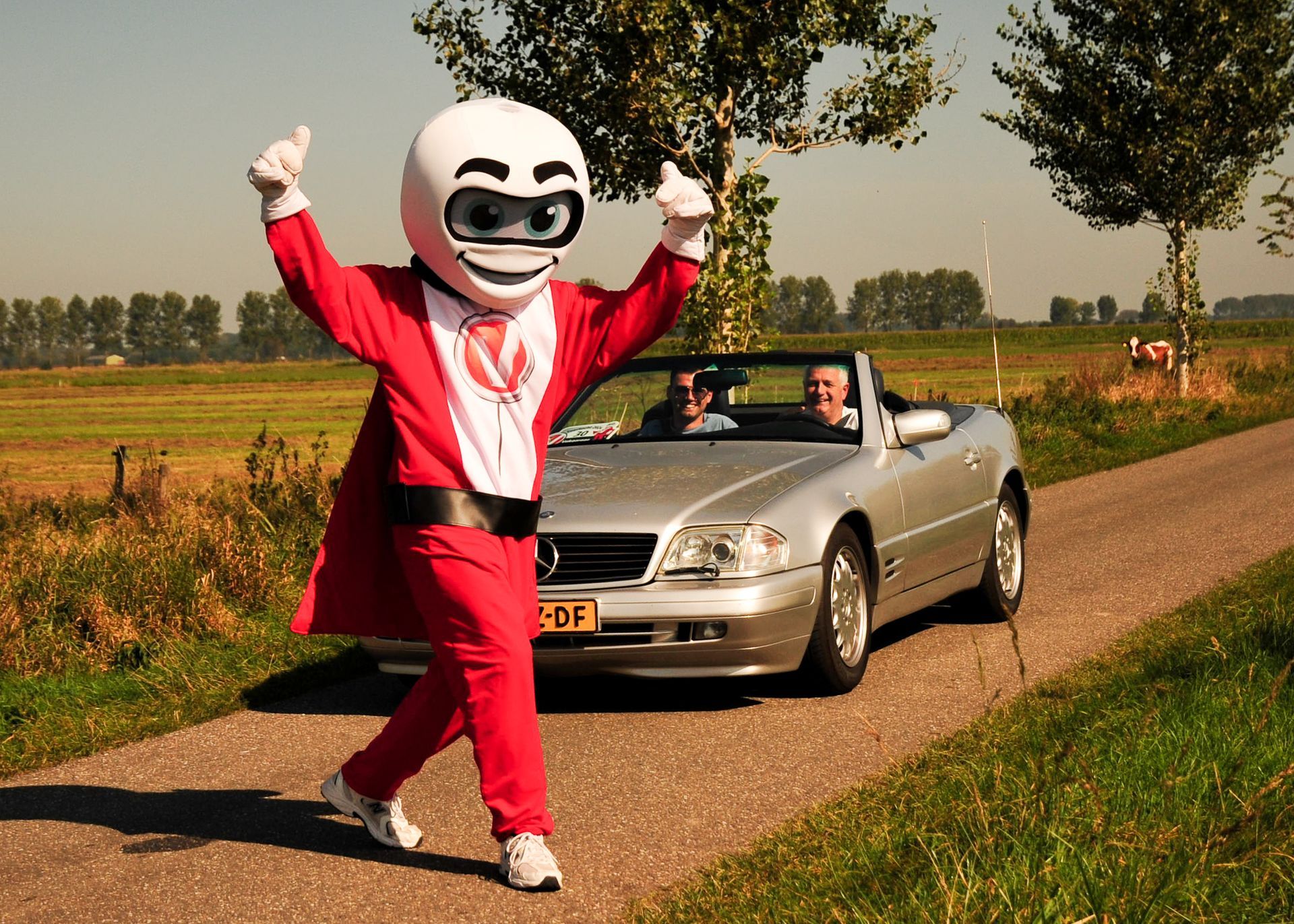 Mascotte in rood pak steekt duim omhoog naast een zilveren cabriolet op de weg.