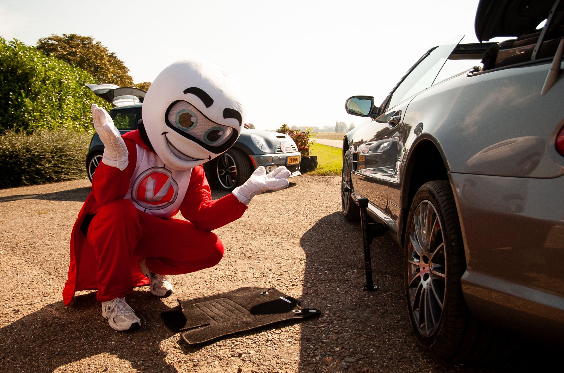 Mascotte knielt naast een auto en gebaart met open handen. Rode overall, helm. Oprit met grind, zonnige dag.
