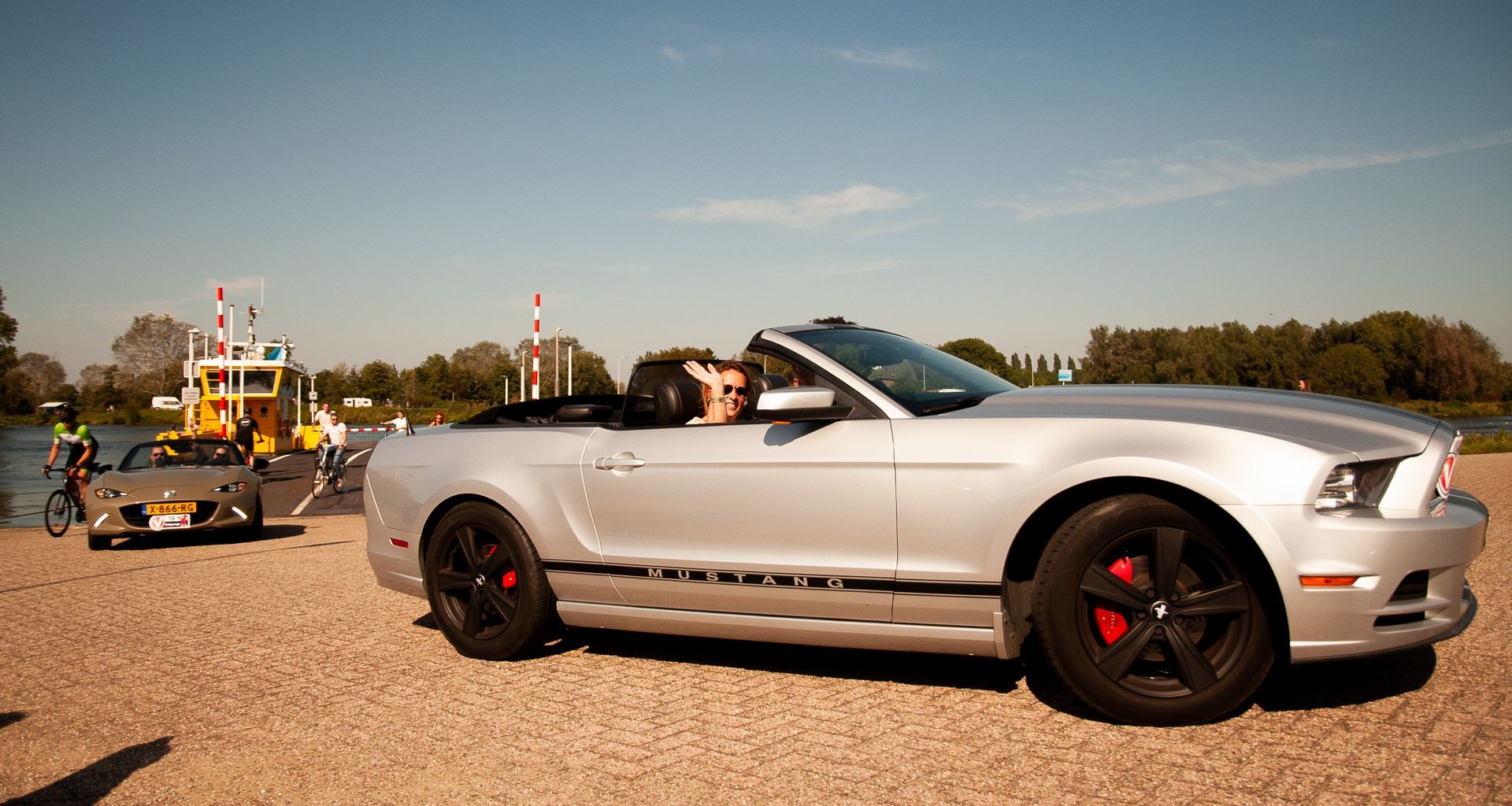 Een zilverkleurige cabriolet Mustang op een kiezelstrand; op de achtergrond zijn nog een auto en een veerboot te zien, het is een zonnige dag.
