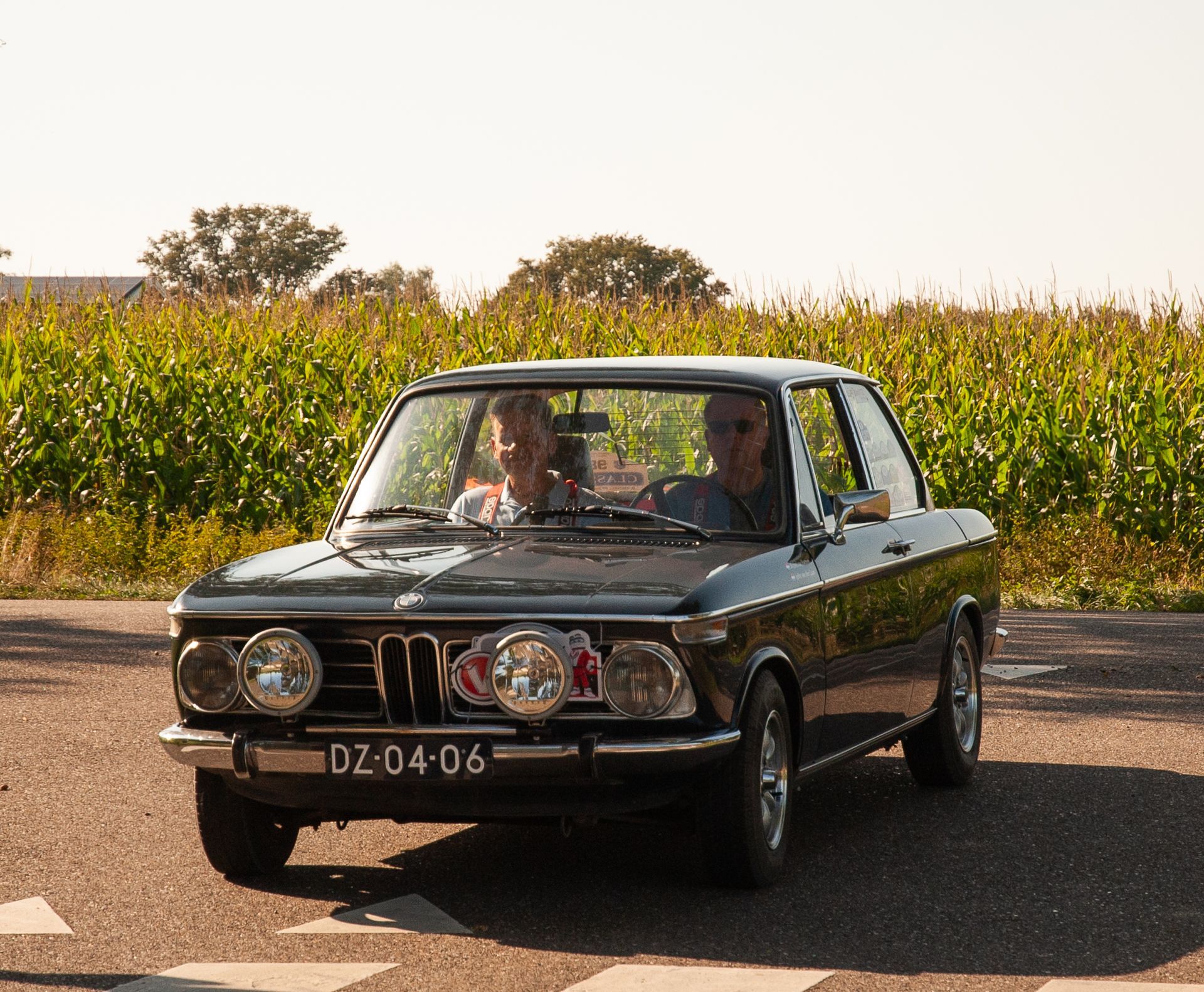 Een zwarte vintage BMW rijdt over de weg, met twee inzittenden en extra koplampen.