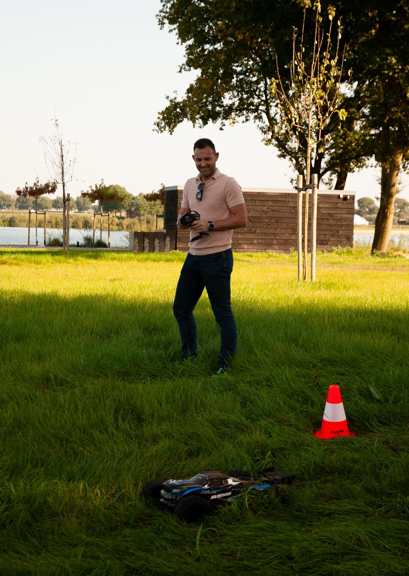 Een man bedient een dronecontroller op een grasveld bij een meer. Een verkeerskegel staat er vlakbij.