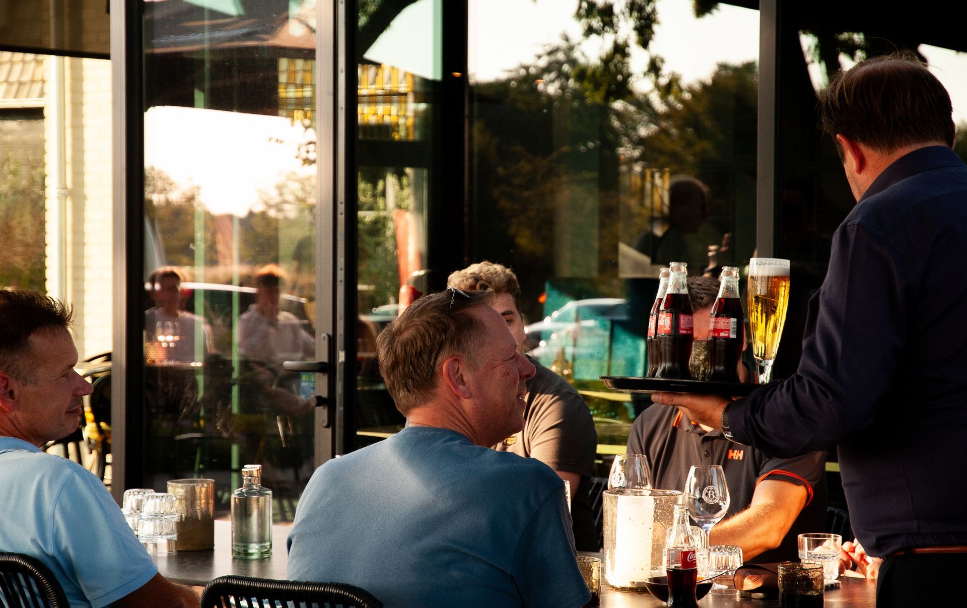 Een ober serveert drankjes in een restaurant met een terras. Mensen zitten aan tafels en worden weerspiegeld in de glazen ramen.